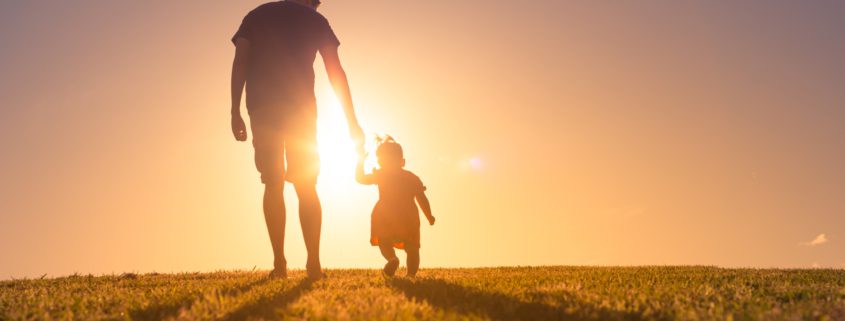 sunset shot of a father with toddler silhouetted agains the sun on a field of low cut grass